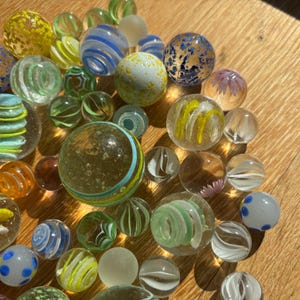 Op de afbeelding: Een verzameling glazen knikkers in verschillende maten en ontwerpen, waaronder doorzichtige bollen met interne wervelingen van geel, blauw, groen en wit, evenals effen, gespikkelde en doorschijnende glazen ballen verspreid over het oppervlak.