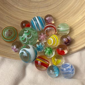 Op de afbeelding: Een verzameling diverse glazen knikkers met verschillende ontwerpen, waaronder bloemmotieven aan de binnenkant, wervelende strepen en effen kleuren in rood, blauw, groen, geel en wit, gepresenteerd in een gebogen houten kom.