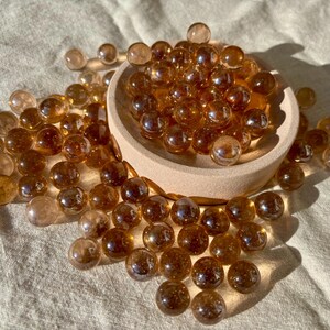 Op de afbeelding: Een verzameling amberkleurige glazen knikkers, sommige verspreid en andere in een kleine, ronde houten kom. De knikkers hebben een reflecterend, glanzend oppervlak en de scène wordt verlicht door natuurlijk zonlicht.