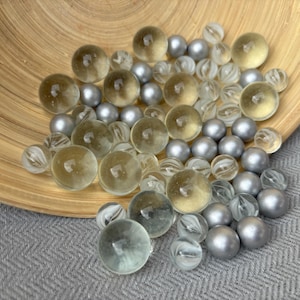 Könnte beinhalten: Eine Sammlung von kugelförmigen Glas- und Metallknöpfen in verschiedenen Größen. Das Sortiment umfasst durchscheinende, gelb getönte Glaskugeln, klare Glaskugeln mit internen Wirbelmustern und massive, matt silberfarbene Metallkugeln.