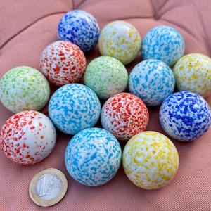 Op de afbeelding: Een verzameling van dertien bolvormige glazen knikkers, elk met een witte basis met gespikkelde patronen in blauw, rood, groen en geel. Een munt van één euro is in de linkerbenedenhoek geplaatst om een duidelijke schaalvergroting te bieden.