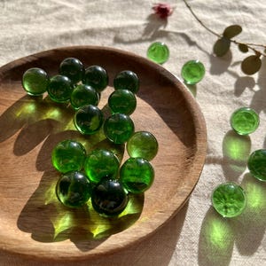 Op de afbeelding: Een houten kom gevuld met groene glazen knikkers. De kom staat op een wit stoffen oppervlak.