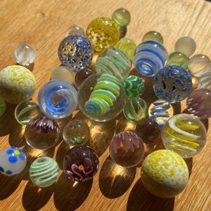 Op de afbeelding: Een verzameling diverse glazen knikkers in verschillende maten, kleuren en patronen. De set bevat heldere, ondoorzichtige en doorschijnende bollen met interne wervelingen, spikkels, bloemachtige ontwerpen en effen kleuren zoals blauw, geel, groen en paars.