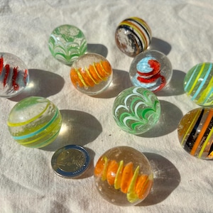 Op de afbeelding: Een verzameling van tien doorzichtige glazen knikkers met diverse interne patronen, waaronder oranje, gele, groene, blauwe, rode en zwarte wervelingen, vergezeld door een tweekeuromunt als schaalvergroting met een diameter van 25,75 mm.