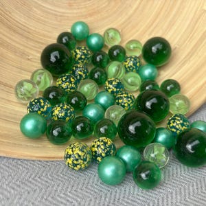Op de afbeelding: Een verzameling glazen knikkers in diverse groentinten, waaronder effen, doorschijnende en heldere varianten. Sommige knikkers hebben gespikkelde gele en groene patronen aan de binnenzijde, terwijl andere een gladde, parelmoerachtige of diepgroene afwerking hebben.