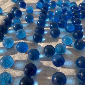 Op de afbeelding: Een verzameling blauwe glazen knikkers verspreid over een wit stoffen oppervlak. De knikkers zijn rond en glad, met een lichte glans.