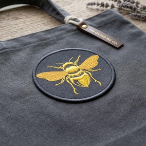 May include: A gray tote bag with a large embroidered patch featuring a golden bee design. The patch is circular and stitched onto the bag. The bag has a leather tag that reads "THE GILDED BEE CO."
