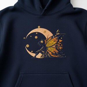 Peut inclure: Sweat à capuche bleu marine avec un motif brodé d'une fée assise sur un croissant de lune. La fée a des ailes orange et jaunes, avec des accents dorés sur la lune et autour de la fée. Le sweat à capuche a une poche avant et une capuche.