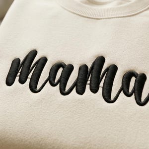 Op de afbeelding: Crèmekleurige sweatshirt met het woord "Mama" in zwarte cursieve letters geborduurd op de borst. De sweatshirt is netjes opgevouwen en de stof voelt zacht aan. De achtergrond is een houten oppervlak.