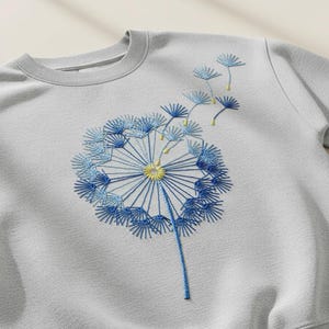 Può includere: Una felpa grigio chiaro presenta un ricamo di un soffione. Il soffione è realizzato in tonalità di blu, con un centro giallo. Il disegno include lo stelo e diversi semi che fluttuano via dal fiore.