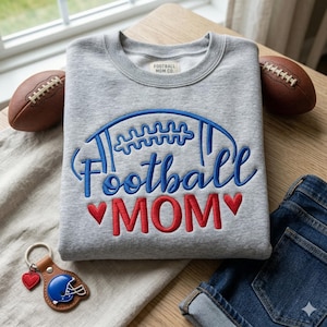 May include: A gray sweatshirt with the words "Football MOM" embroidered in blue and red. The sweatshirt has a football design above the text. A brown leather keychain with a blue football helmet and a red heart is next to the sweatshirt.