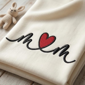 Könnte beinhalten: Cremefarbene Decke mit dem gestickten Wort "Mom" in schwarzer Schrift, wobei ein rotes Herz das "o" ersetzt. Ein kleines, beiges Häschen-Spielzeug aus Strick befindet sich im Hintergrund.
