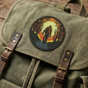 May include: Olive green canvas backpack with brown leather straps and a circular embroidered patch. The patch features a silhouetted figure walking towards a sunset in a forest setting, with orange, yellow, and black colors.