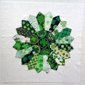 May include: A circular quilt block featuring a shamrock design. The block is composed of various green fabrics with shamrock patterns, arranged in a radial pattern against a white background. The design celebrates St. Patrick's Day.