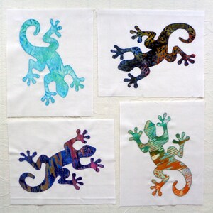 May include: Four fabric squares each feature a colorful lizard applique. The lizards are in shades of blue, green, orange, and purple, with intricate patterns. The appliques are stitched onto white fabric squares.