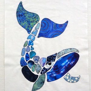 May include: A fabric art piece featuring a whale design. The whale is composed of various blue fabric pieces with different patterns, including swirls and floral prints. The background is plain white.