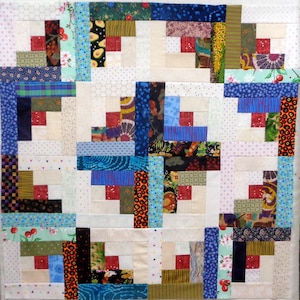 May include: A handmade quilt square featuring a log cabin design. The square is composed of various fabric scraps in a patchwork style, with a central red square surrounded by rectangular strips of different patterned fabrics, including floral, polka dot, and geometric designs.