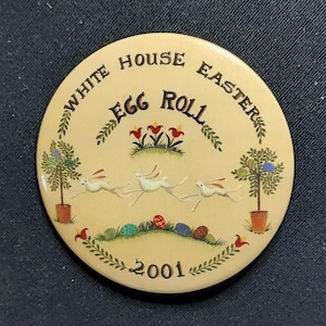 Puede incluir: Un pin redondo de color crema con el texto "WHITE HOUSE EASTER EGG ROLL 2001". El pin presenta ilustraciones de conejos blancos corriendo, huevos de Pascua coloridos y árboles en macetas. Elementos florales decorativos rodean el texto y las imágenes.