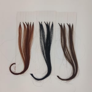 May include: Three hair extensions in varying shades of brown and black are displayed against a white background. Each bundle of hair is attached to a small white card with a series of fine threads. The hair colors range from light brown to dark black.