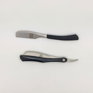 May include: Two straight razors with silver blades and black handles are displayed against a white background. The text "23 THE LUCKY EDITION" is printed on the blade of the top razor. The bottom razor has the number "23" printed on the blade.