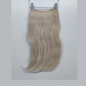 May include: A long, wavy, light blonde hair extension. The hair is hanging from a metal clip.