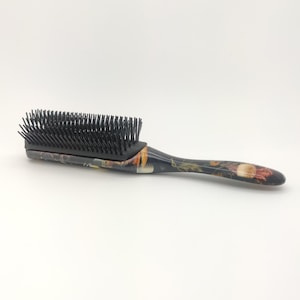 Op de afbeelding: Een haarborstel met een zwart borstelkussen en een handvat met patroon. De handgreep heeft een mix van kleuren, waaronder bruin, oranje en wit. De borstel is ontworpen voor het ontwarren en stylen van haar.