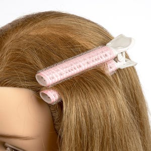 Puede incluir: Primer plano de una cabeza de maniquí con cabello castaño claro peinado con dos rulos de velcro rosa y blanco asegurados con pinzas de plástico blanco. Los rulos son cilíndricos y tienen una superficie suave y texturizada.