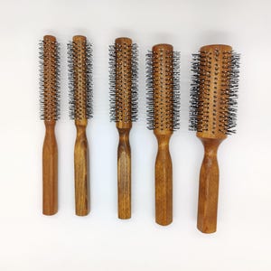 Puede incluir: Cinco cepillos redondos de madera para el cabello con cerdas metálicas sobre un fondo blanco. Los cepillos varían en tamaño, con mangos de madera marrón claro y cabezales cilíndricos. Las cerdas son plateadas y densas.