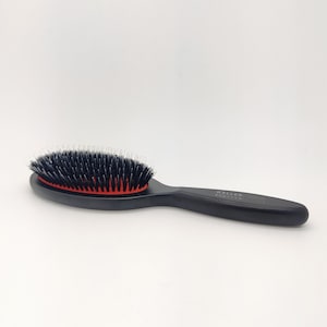 Puede incluir: Cepillo de pelo ovalado negro con cerdas rojas y un mango negro. El mango tiene el texto "KELLER PROFESSIONAL" impreso.