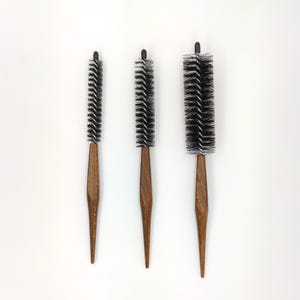 Puede incluir: Tres cepillos redondos para el cabello con mangos de madera y cerdas negras se disponen sobre un fondo blanco. Los cepillos varían en tamaño, con cerdas densas y mangos puntiagudos. Los mangos de madera tienen un tono cálido y natural.