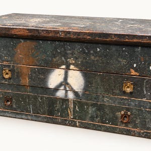 May include: A rectangular wooden chest with a distressed dark green finish, featuring a white spray-painted peace sign on the front. The chest has two horizontal drawers with small, recessed brass-toned metal pulls and a flat, hinged lid top.