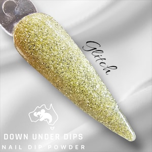 Może przedstawiać: Zbliżenie na złoty brokatowy puder do zanurzania paznokci z napisem "Glitch" napisanym czarnym kursywą na białym tle. Tekst "Down Under Dips Nail Dip Powder" jest również widoczny na dole obrazu.