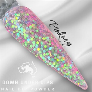 May include: Pinky nail dip powder with a holographic glitter finish. The powder is in a clear container with a pink and white label that says "Down Under Dips Nail Dip Powder".