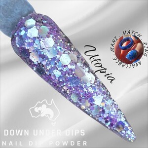 May include: A nail dip powder sample with a glittery, iridescent finish. The sample is a gradient of purple and blue, with large, hexagonal glitter pieces. The text "Utopia" and "Down Under Dips Nail Dip Powder" are visible.