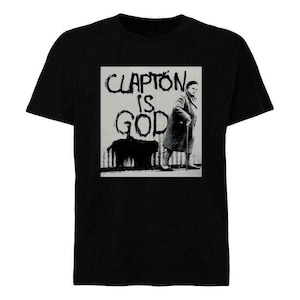 Puede incluir: Camiseta negra con un diseño gráfico. El diseño presenta el texto "CLAPTON IS GOD" en una fuente desgastada, junto con una imagen en blanco y negro de una persona con un bastón. Una camiseta de inspiración rock and roll clásica.