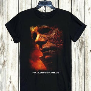 Könnte beinhalten: Schwarzes T-Shirt mit einer Nahaufnahme eines verwitterten Gesichts mit Maske in Orange- und Brauntönen. Der Text "HALLOWEEN KILLS" ist unten in Weiß aufgedruckt. Das T-Shirt hängt an einem schwarzen Kleiderbügel.