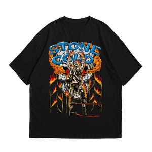 May include: Black t-shirt featuring a graphic of a wrestler with the text "STONE COLD" above a flaming skull. The wrestler is in a wrestling ring, with flames around the edges. The design is in shades of orange, blue, and white.