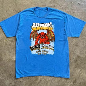 May include: A blue t-shirt featuring a graphic of a red devil with bat wings holding a baseball bat. The shirt has the word "SHURIK" in yellow and orange, and the words "INGROWN FORENA" and "GOD HATE JOE BIE" in a blue font.
