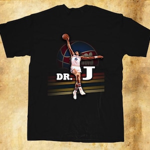 May include: Black t-shirt featuring a vintage graphic of a basketball player mid-air, holding a basketball. The design includes the text "DR. J" and a retro color scheme. The player is wearing a white uniform with the number 6.