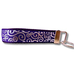 May include: A purple key fob wristlet with a silver swirl pattern. The key fob has a gold-coloured metal clasp and key ring. The wristlet is approximately 15 cm long.