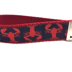 May include: A red key fob with a navy blue band featuring a repeating pattern of red lobster illustrations. The key fob has a silver-colored metal clasp for attaching keys. The key fob is made of a woven fabric.