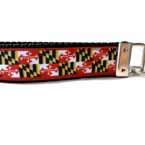 May include: A key fob featuring the Maryland state flag design. The fob has a black border and a red, white, yellow, and black pattern. It includes a silver-colored metal key ring and hardware.