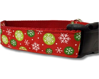 May include: A red pet collar featuring a repeating pattern of white snowflakes and green circular ornaments. The collar is constructed from a textured fabric and includes a black plastic side-release buckle for secure fastening.