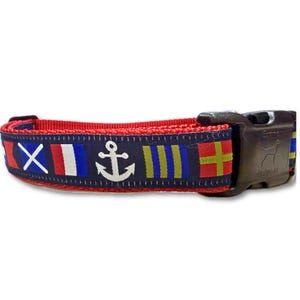 Op de afbeelding: Een rode en marineblauwe hondenriem met een nautisch vlaggenontwerp. De halsband heeft een witte anker en verschillende vlag symbolen. De gesp is zwart en heeft een honden silhouet.