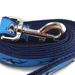 May include: A blue and navy dog leash with a silver clip. The leash has a repeating pattern of white sharks with blue and white water splashes.