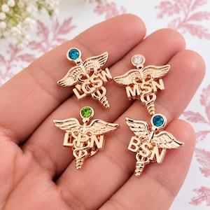 Puede incluir: Cuatro broches m&eacute;dicos de caduceo en tono oro rosa con peque&ntilde;as piedras de estr&aacute;s en la parte superior. Cada broche muestra un acr&oacute;nimo diferente: LCSW con piedra azul, MSN con piedra clara, LPN con piedra verde y BSN con piedra azul.