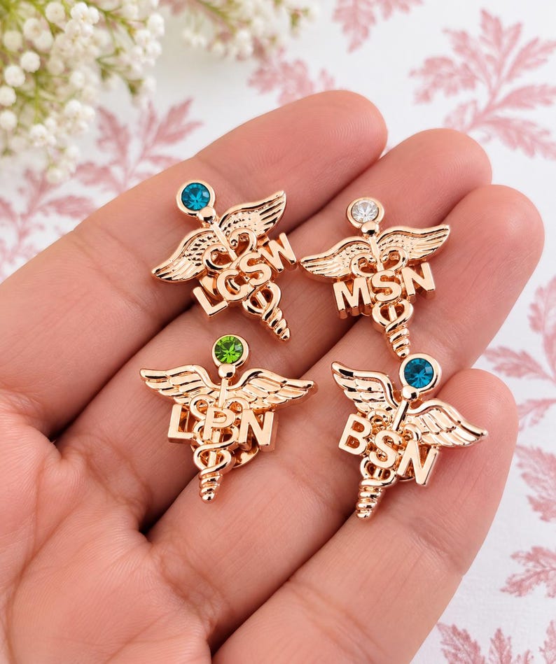 Puede incluir: Cuatro broches m&eacute;dicos de caduceo en tono oro rosa con peque&ntilde;as piedras de estr&aacute;s en la parte superior. Cada broche muestra un acr&oacute;nimo diferente: LCSW con piedra azul, MSN con piedra clara, LPN con piedra verde y BSN con piedra azul.