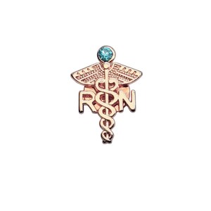 Puede incluir: Un broche de metal en tono oro rosa que presenta un s&iacute;mbolo de caduceo con las letras RSN integradas en el dise&ntilde;o. La parte superior del broche est&aacute; acentuada con una piedra preciosa redonda de color azul claro engastada en el centro de las alas.