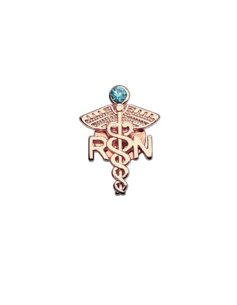 Puede incluir: Un broche de metal en tono oro rosa que presenta un s&iacute;mbolo de caduceo con las letras RSN integradas en el dise&ntilde;o. La parte superior del broche est&aacute; acentuada con una piedra preciosa redonda de color azul claro engastada en el centro de las alas.