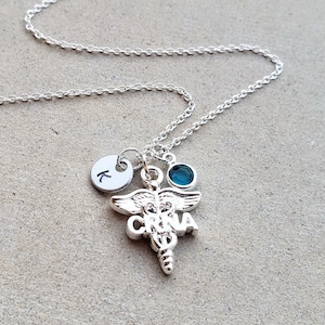 May include: Silver necklace with a charm of a medical caduceus with the letters "CRNA" on it. There is also a small silver charm with the letter "K" on it and a teal blue gemstone.